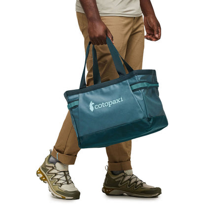 Person carrying a teal Cotopaxi Allpa 30L Gear Hauler Tote, walking in khaki pants and hiking shoes.
