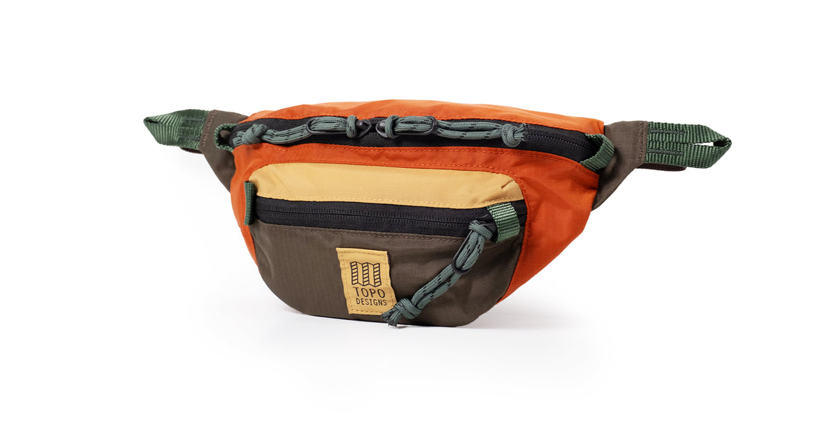 Topo Designs Mountain Waist Pack in orange, tan, brown, and green with zippers and cord detailing on a white background.
