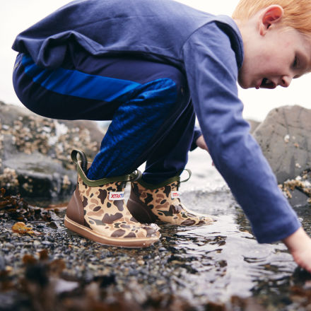 Child in XTRATUF Kids' Ankle Deck Boots - Stone, crouching and exploring a rocky tide pool.