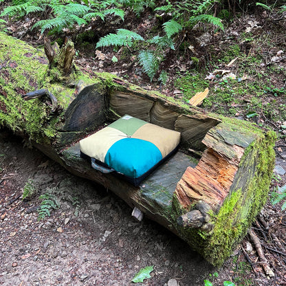 Wanderer 'QUAD' Outdoor Cushion by Media Mercantile, featuring green, tan, and teal panels, rests on a mossy log in the forest.