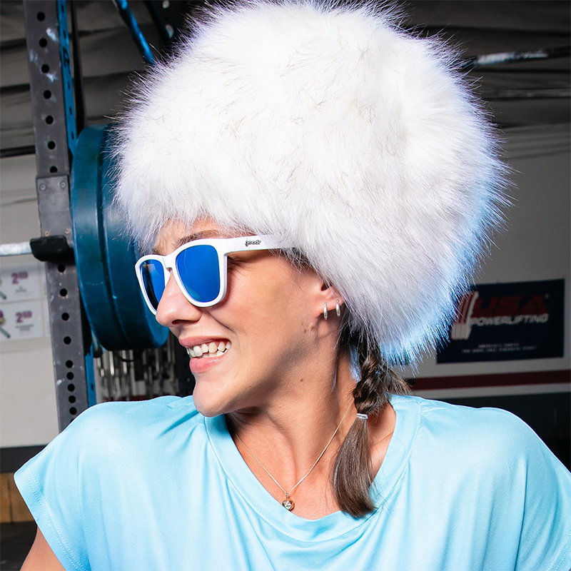 A person wearing white Iced by Yetis goodr sunglasses with blue reflective lenses and a fluffy white hat, smiling in a gym.