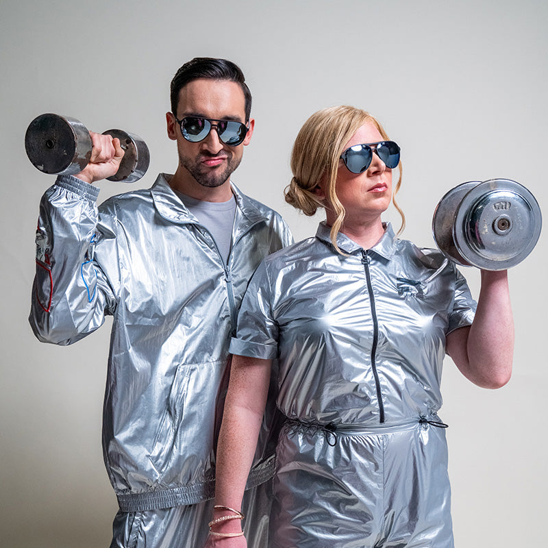 Two athletes in metallic silver outfits and goodr 'Add the Chrome Package' mirrored sunglasses, posing with dumbbells.