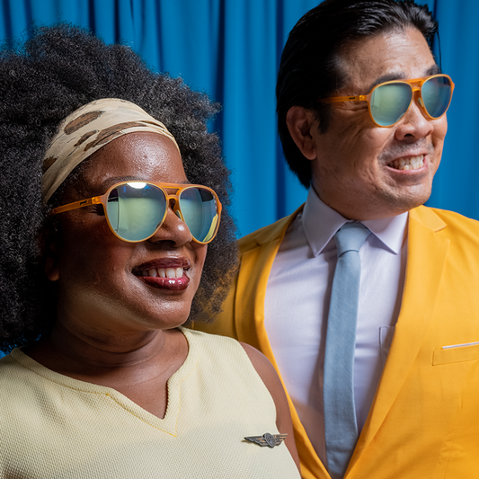 goodr Cheesy Flight Attendant sunglasses on a smiling Black woman in a yellow top and an Asian man in a yellow suit.