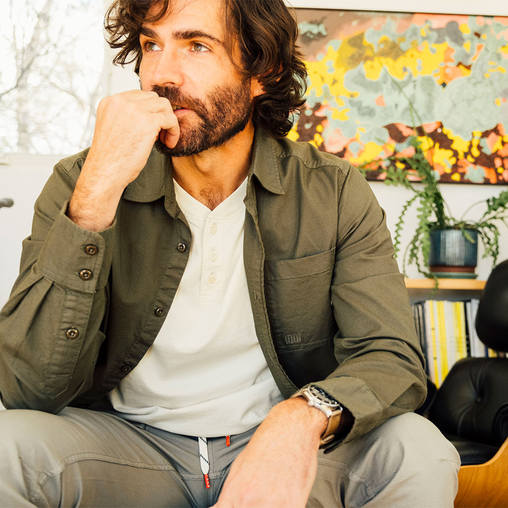 Man wearing a green Topo Designs Men's Desert Shirt over a henley, with gray pants, in a casual indoor setting, looking thoughtful.