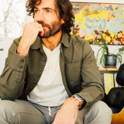 Man wearing a green Topo Designs Men's Desert Shirt over a henley, with gray pants, in a casual indoor setting, looking thoughtful.