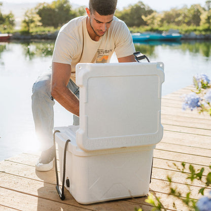Man on a sunny dock by a lake opening a white 25 QT Bison Cooler from Bison Coolers, ready for outdoor adventures.