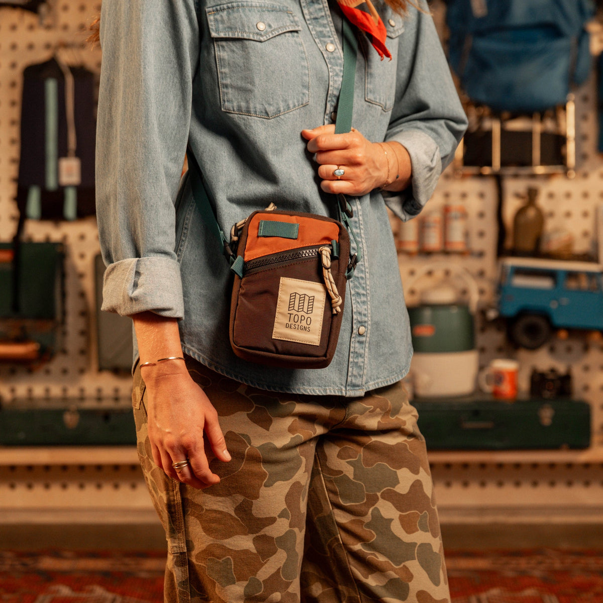 Topo Designs Mini Shoulder Bag in brown and orange, worn by a person in a denim shirt and camo pants.