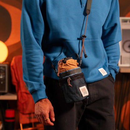 Mountain Hydro Sling by Topo Designs worn by a person in a blue hoodie, showcasing the black mesh bottle holder with brown top.