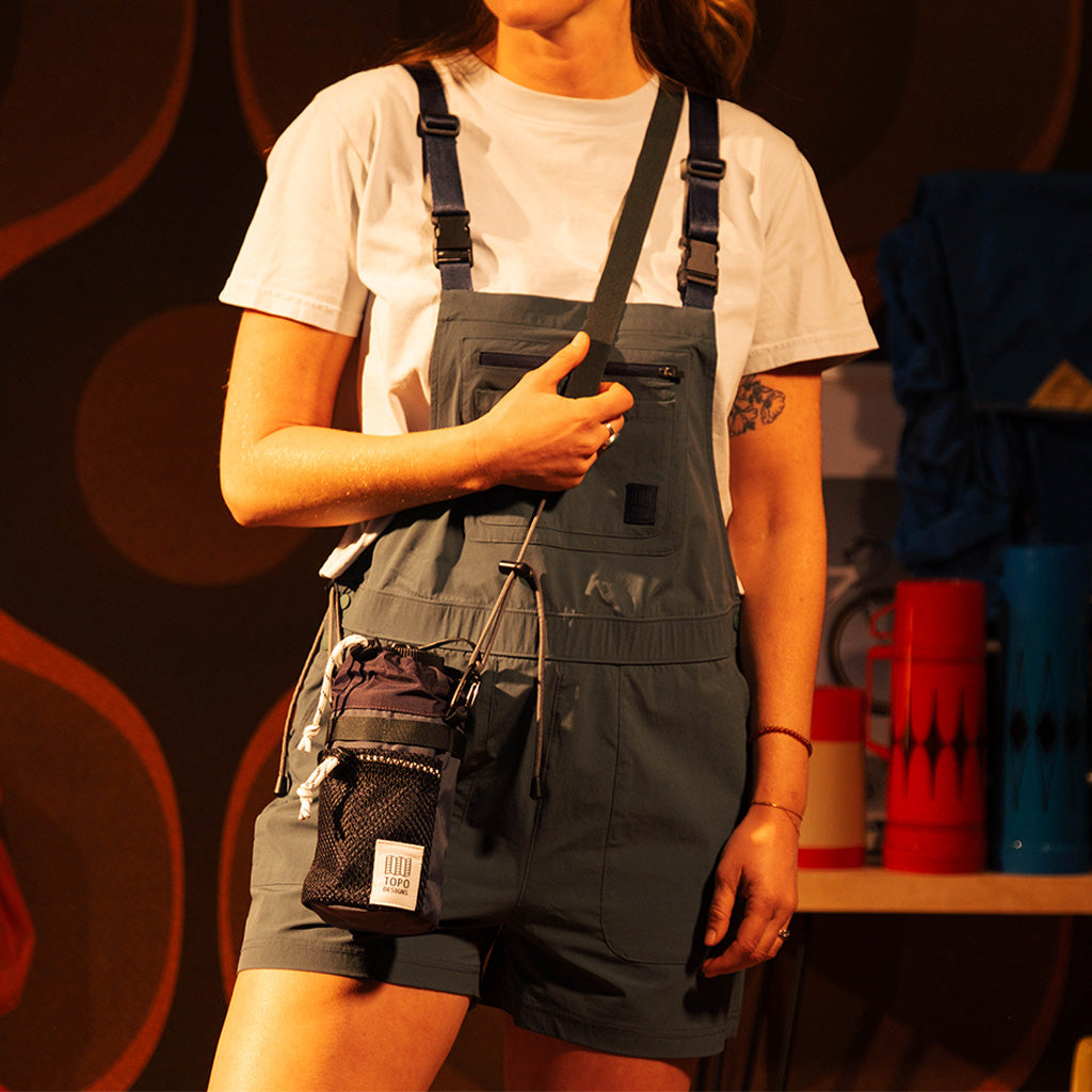 Person wearing grey overalls and a white t-shirt carrying a dark grey Topo Designs Mountain Hydro Sling with a bottle.
