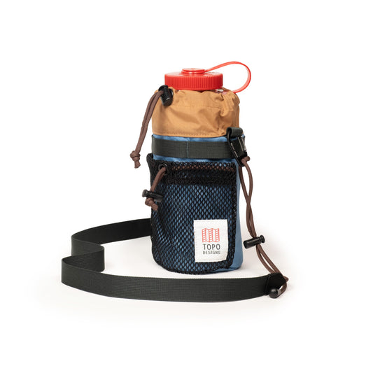 Topo Designs Mountain Hydro Sling in blue and brown, holding a water bottle with a red cap, featuring a mesh pocket and black adjustable strap.