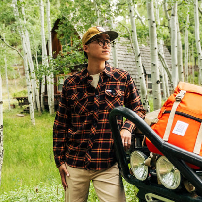 Man wearing a Topo Designs Men's Mountain Shirt Long Sleeve (black/orange plaid), khaki pants, and a cap, in a forest.
