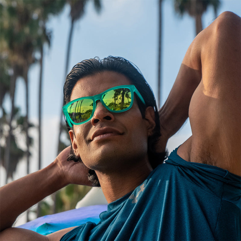 Man in green mirrored goodr Nessy's Midnight Orgy sunglasses, relaxing outdoors with palm trees reflecting in the lenses.