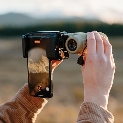 Hands using Nocs Provisions Photo Rig Smartphone Adapter on a monocular, capturing a distant natural landscape on phone screen.