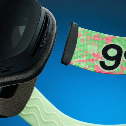 Close-up of goodr Polar Pistachio goggles. Black frame, grey foam, lime green strap with pink splatter design and '9' detail.