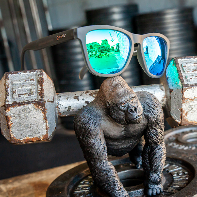 Gray goodr Silverback Squat Mobility sunglasses on a gorilla figurine, surrounded by rusted gym weights. Lenses reflect vehicles.