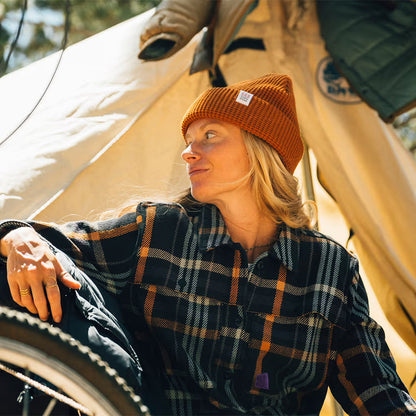 Woman wearing an orange Topo Designs Waffle Knit Beanie and a plaid shirt, sitting inside a tent with a bicycle.